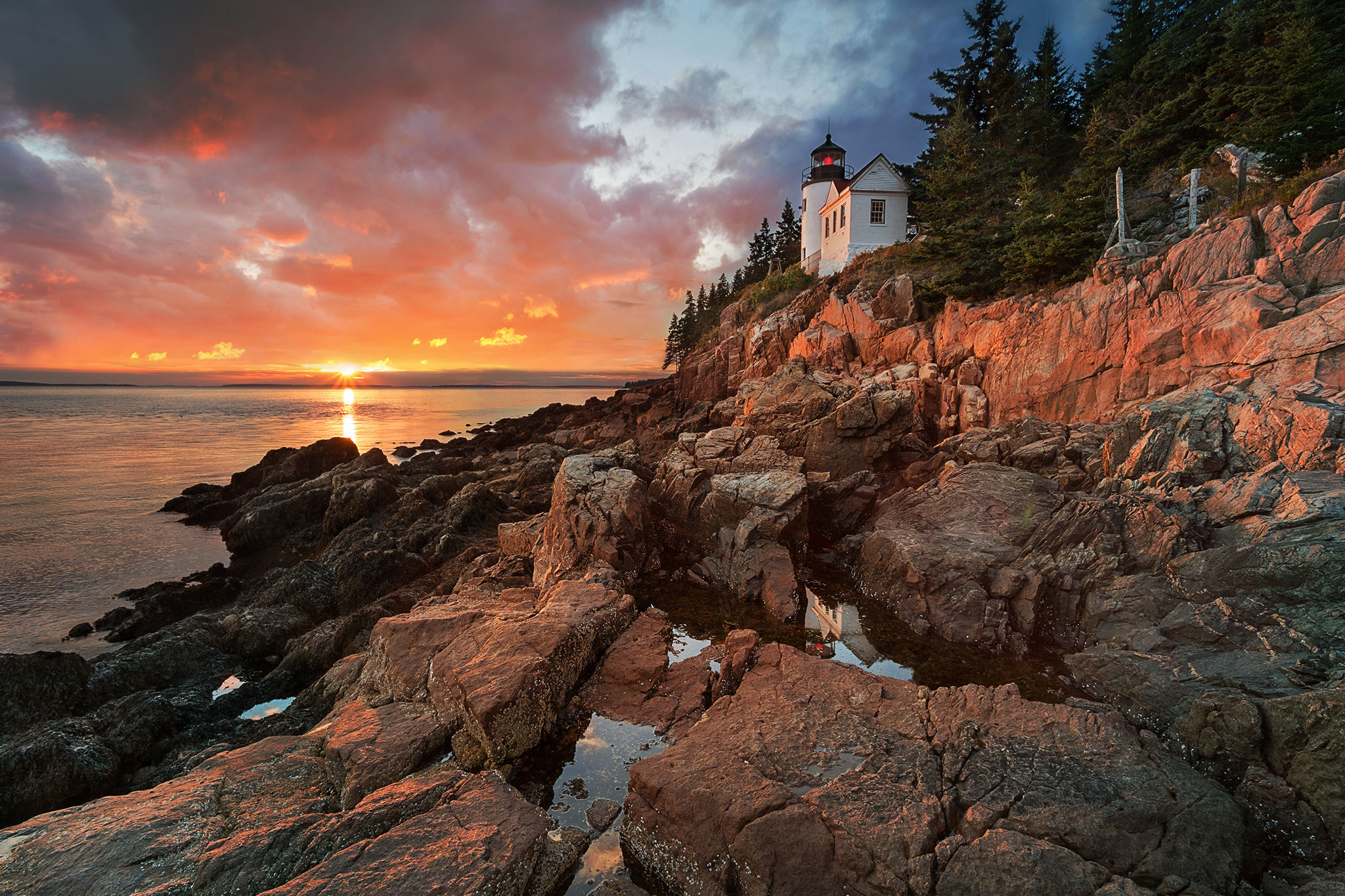 A serene sunset over a calm sea, casting a warm glow on the clouds and illuminating a small white lighthouse perched atop rugged rocky cliffs surrounded by pine trees.