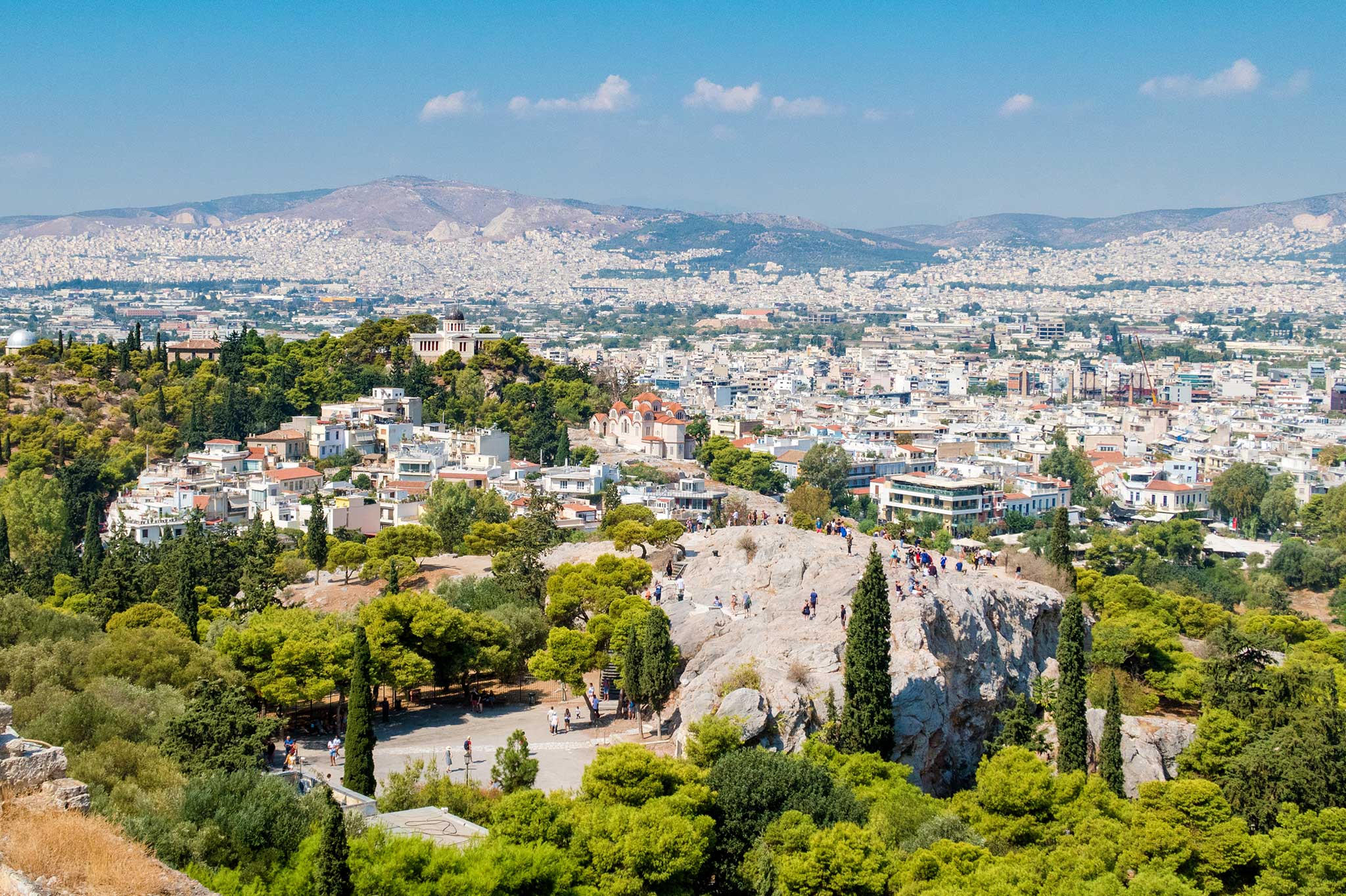 Panoramic view of Athens with dense white buildings, greenery, and mountains in the background, seen from the rocky Mars Hill.