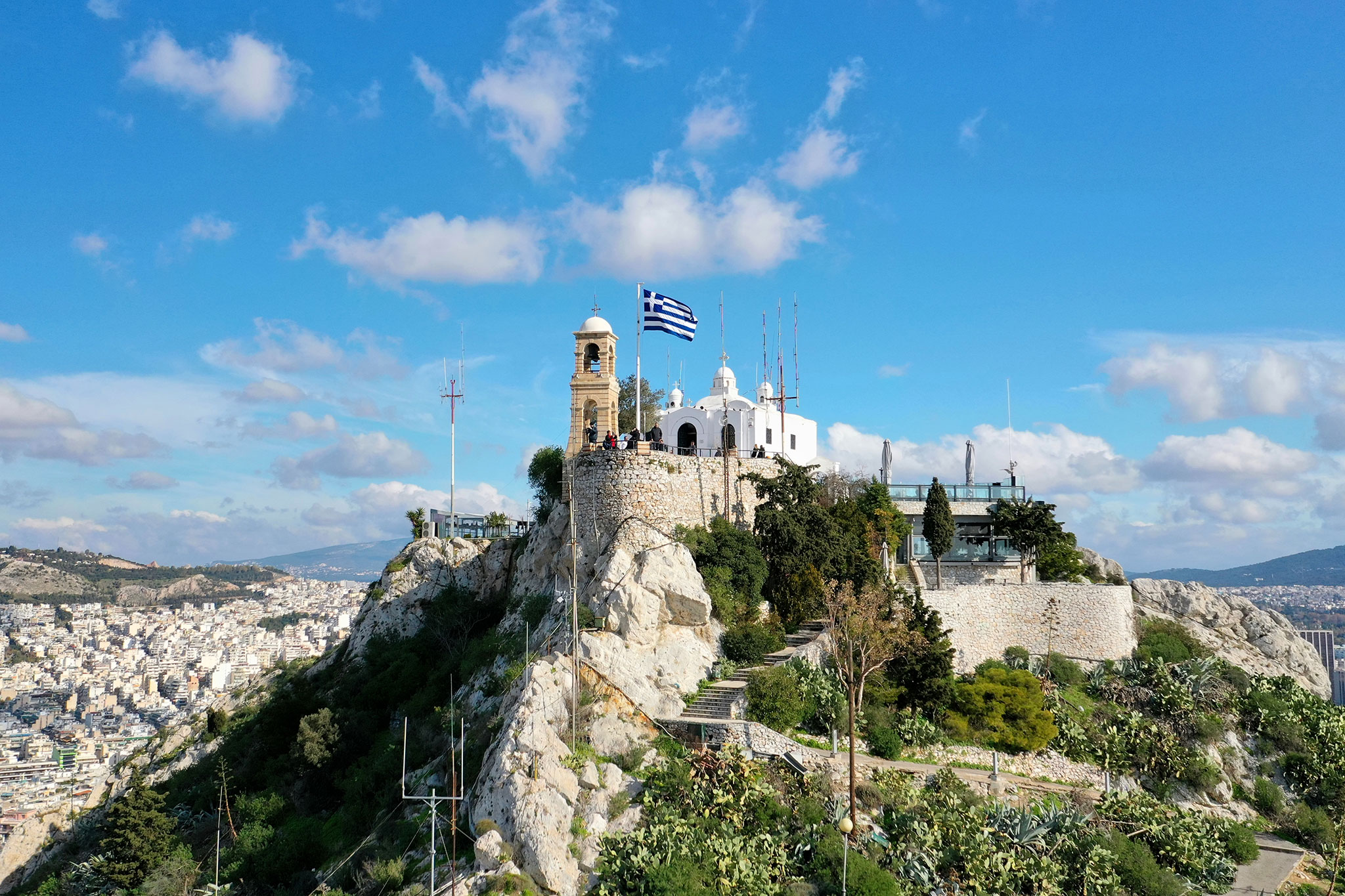 Aerial view of the Chapel of Saint George atop Lycabettus Hill in Athens, with a panoramic view of the city and mountains in the distance under a cloudy sky.
