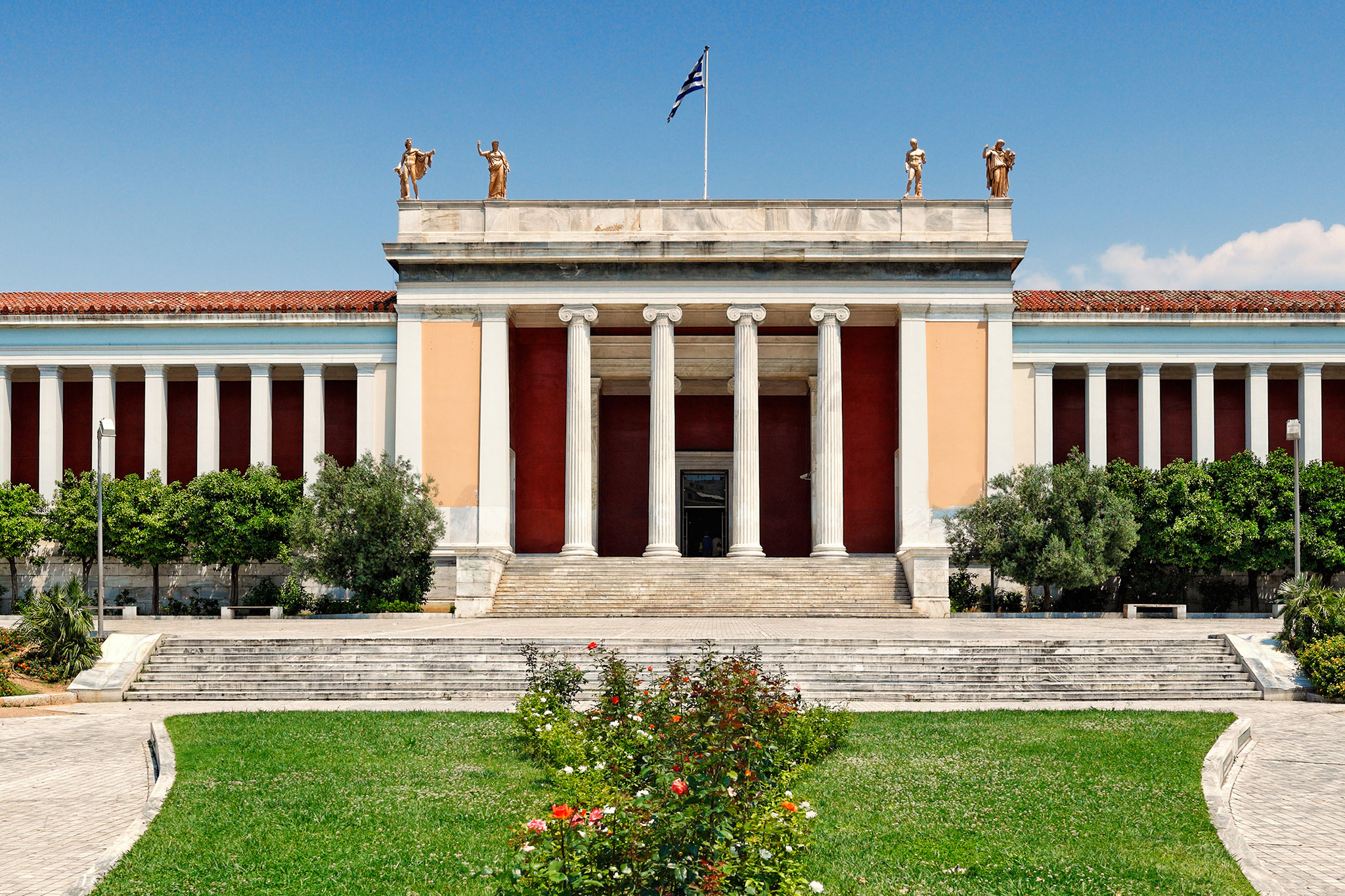 The National Archaeological Museum in Athens, Greece, has classical columns, red walls, a Greek flag, and statues atop the entrance under a blue sky.