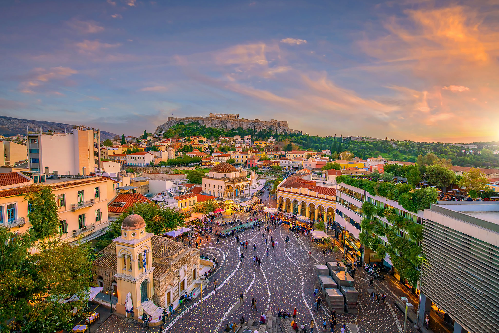 Sunset over Athens, showcasing the city’s skyline with the Acropolis in the background and the lively Monastiraki Square filled with people.