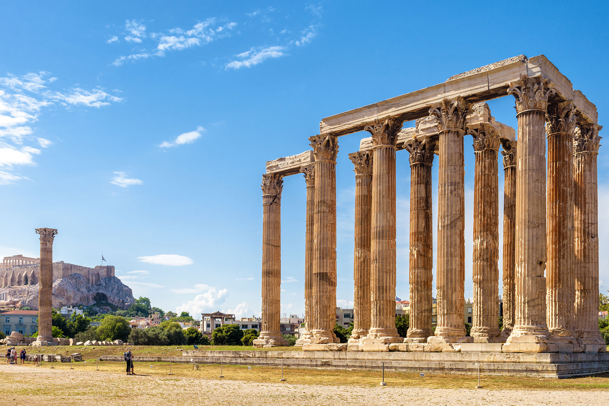 The ancient Greek Temple of Olympian Zeus, with its remaining Corinthian columns standing tall and the Acropolis visible on the hilltop in the distance under scattered clouds.