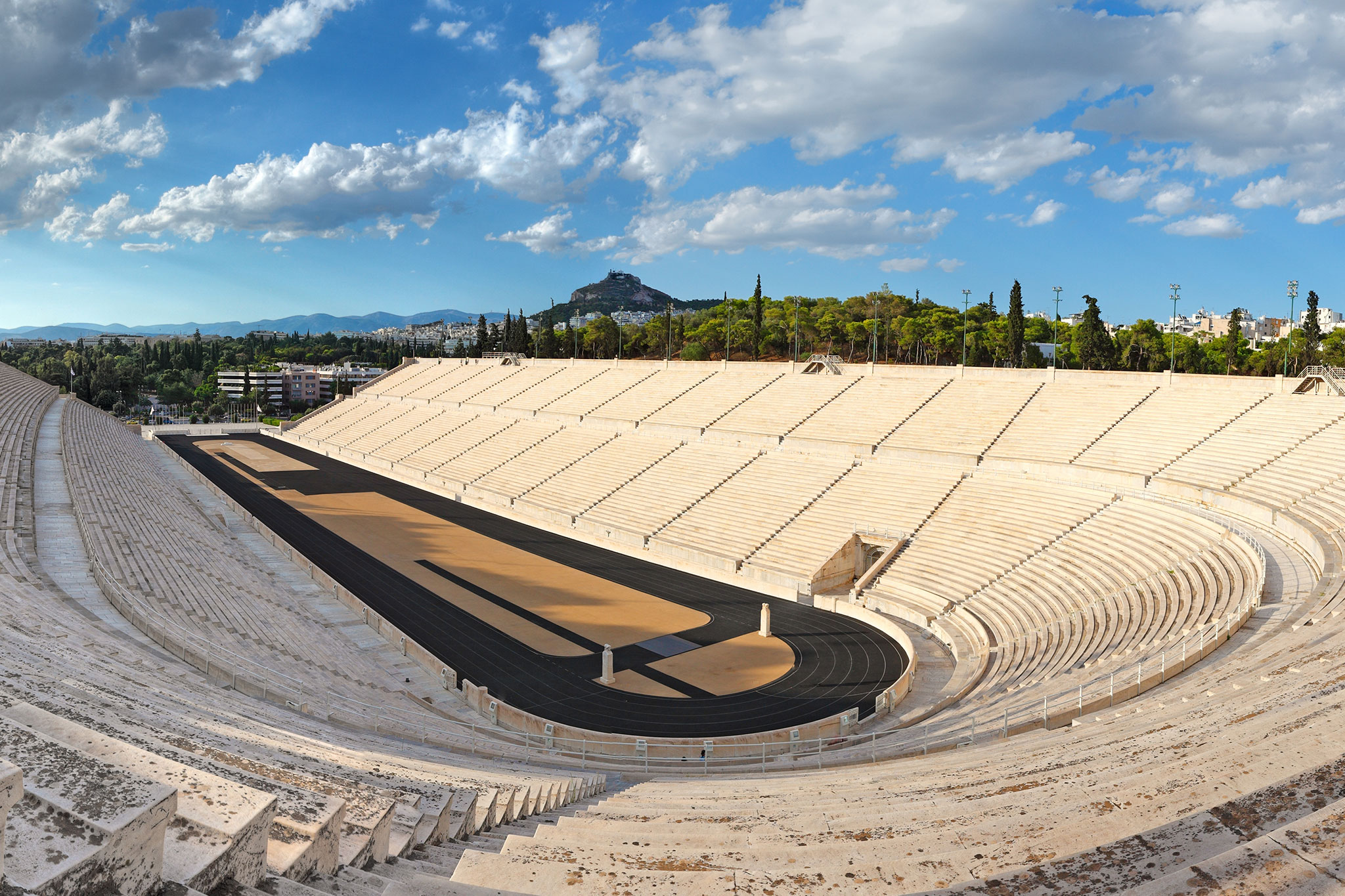 Wide-angle view of the Panathenaic Stadium in Athens, with its distinctive U-shape structure of white marble seats and a black running track under clear blue skies.