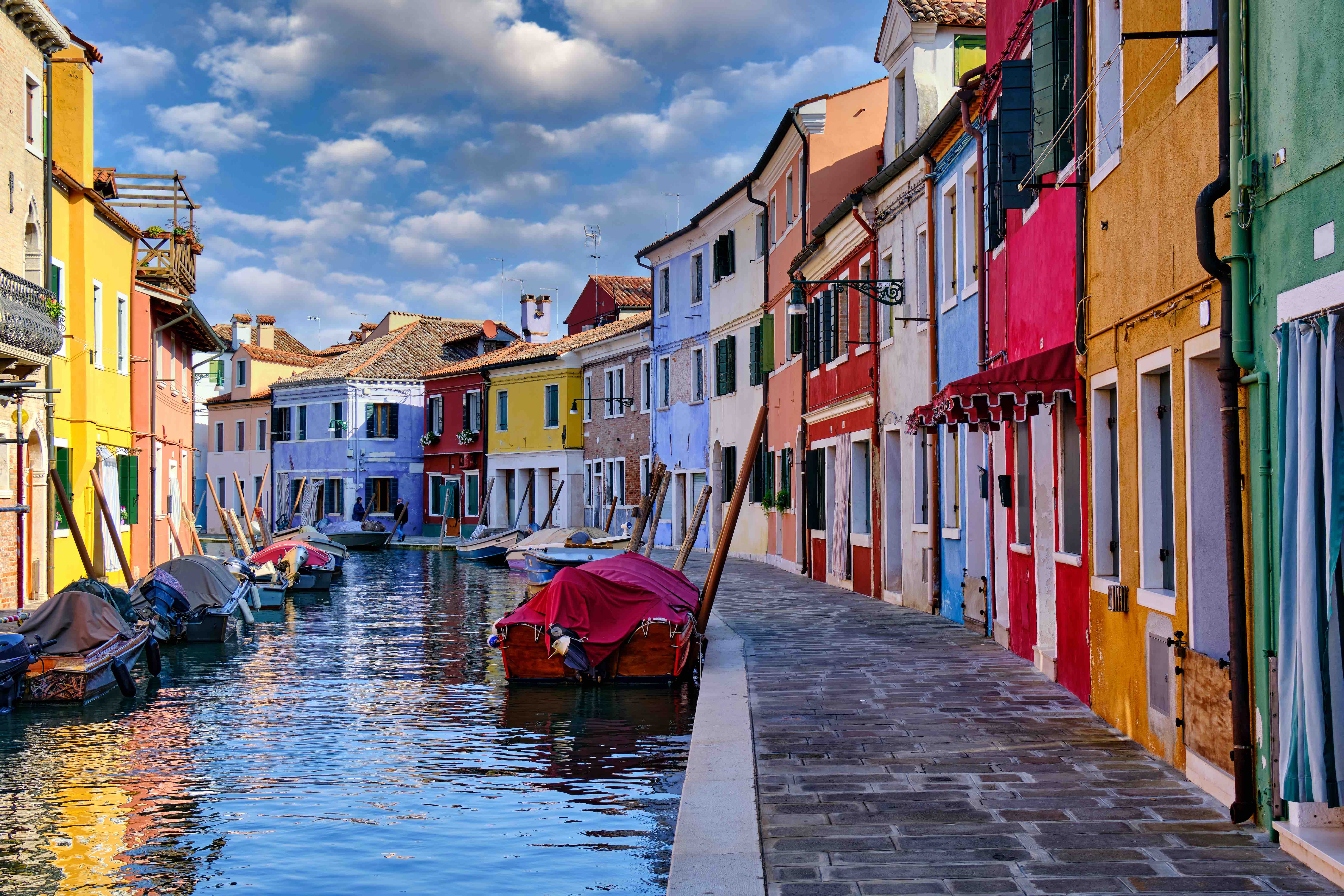 Colorful buildings and boats along a canal and sidewalk in Burano, Italy