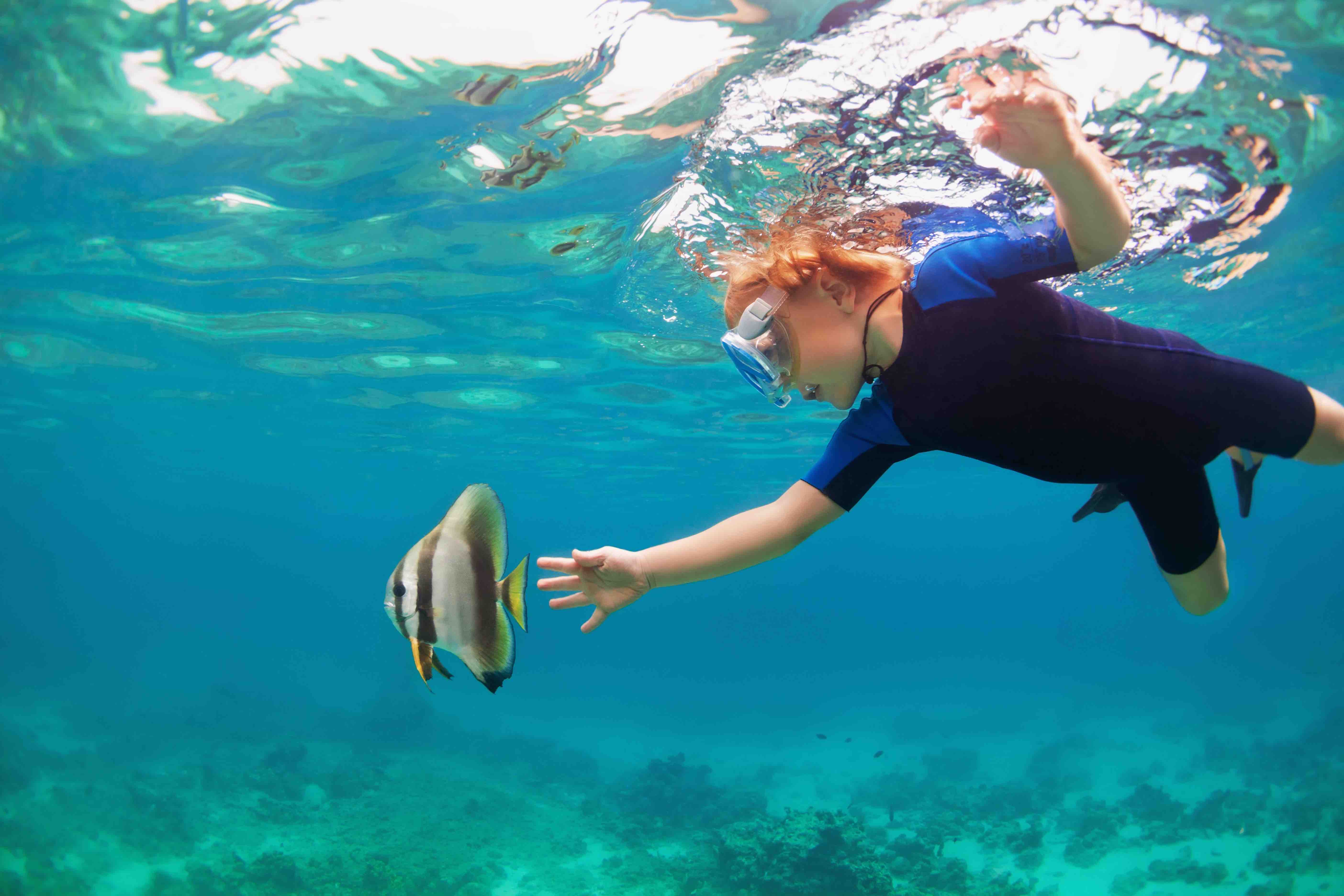 Young child in a snorkeling mask, swimming underwater and reaching for a tropical fish